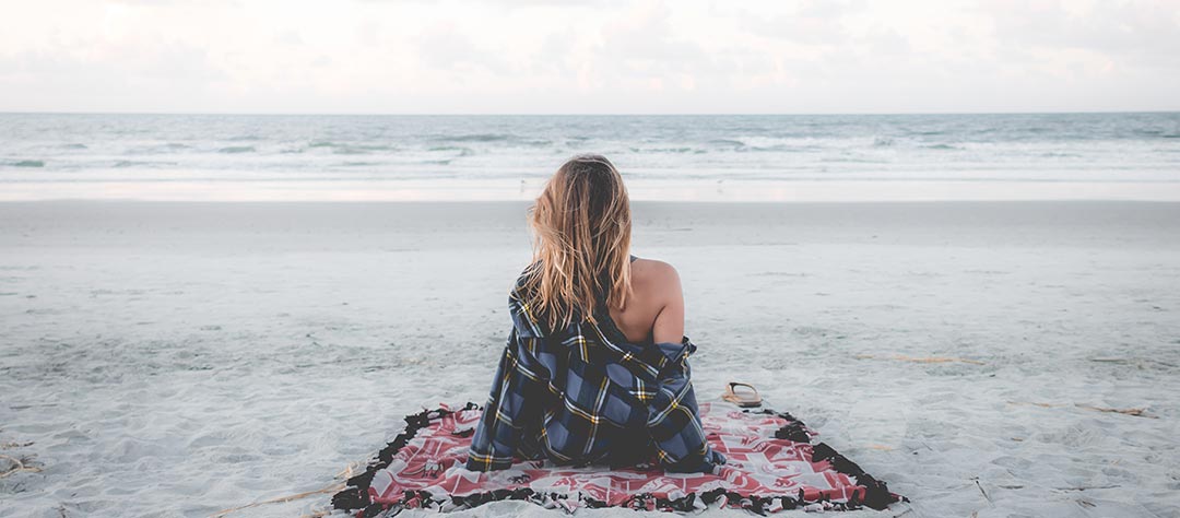 Femme de dos assise sur la plage face à la mer